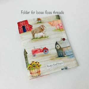 May include: A red, white, and blue fabric folder with a farm theme. The folder has a red barn, a tractor, a pig, a goat, a sunflower, and a farmhouse. The text "Kreative Card Designs" is printed on the folder.