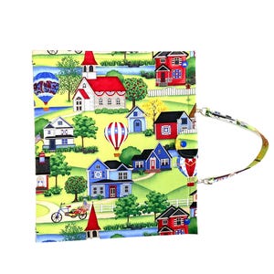May include: A fabric case with a colorful village scene featuring houses, hot air balloons, and a church. The case has a button closure and a patterned strap. The background is yellow.