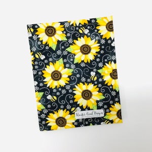 May include: Black fabric with a repeating pattern of yellow sunflowers, white flowers, and bees. The fabric has a white label with the text "Kreativ Card Designs".