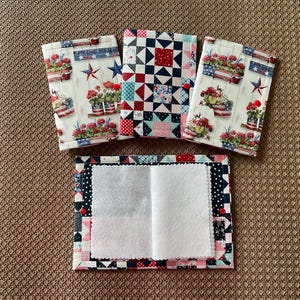 May include: Four fabric items with patriotic designs. Three are small, rectangular, and patterned with floral arrangements, stars, and stripes. The fourth is open, revealing a white interior. The items are arranged on a brown textured surface.
