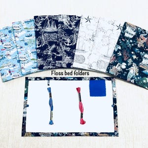May include: A selection of floss bed folders with fabric covers featuring nautical and mermaid designs. The folders surround a white board with blue and pink embroidery threads and a blue fabric square. The text "Floss bed folders" is displayed.