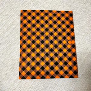 May include: A rectangular fabric folder featuring a vibrant orange and black diagonal gingham check pattern. The folder is secured with a single matching orange snap button closure positioned on the right side, with visible stitching along the edges.