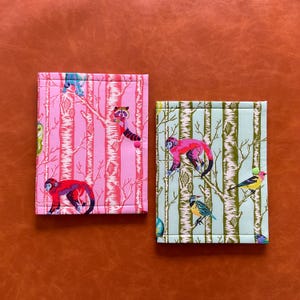 May include: Two fabric-covered notebooks. One is pink with a tree and animal print, featuring red monkeys and a raccoon. The other is light green with a similar tree and animal print, with pink monkeys and colorful birds.