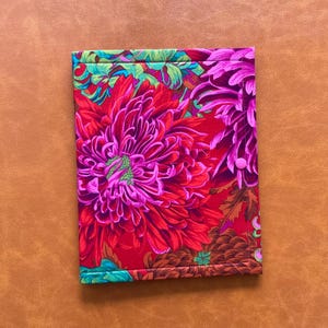 May include: A floral fabric case featuring a vibrant design of large red and pink flowers with green and blue accents. The case is secured with a pink button closure and is set against a brown background.