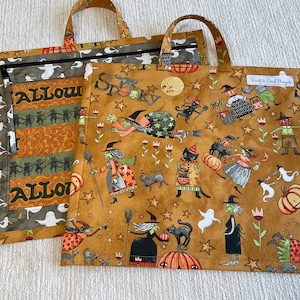 May include: Two square fabric tote bags with handles featuring a Halloween print on a mustard yellow background. The pattern includes witches, black cats, ghosts, pumpkins, and stars. One bag has a zip closure and the text Halloween, while the other displays the word Spooky and a small white label.