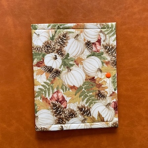 May include: A rectangular fabric item featuring a fall design with white pumpkins, pine cones, and autumn leaves in green, orange, and brown. An orange button secures the item. The background is a solid orange.