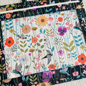 May include: A rectangular fabric pouch featuring a clear vinyl window. The fabric has a dark background with a colorful botanical print of flowers, stems, and birds. A white zipper runs along the top edge of the pouch, securing the contents inside.