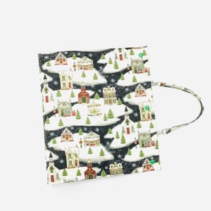 May include: A fabric tote bag featuring a winter village scene with snow-covered houses, trees, and snowflakes on a black background. The bag has a patterned strap and is likely used for carrying items.