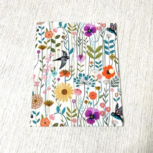 May include: A rectangular fabric pouch featuring a white background with a colorful botanical print of various flowers, green stems, and black birds in flight. The pouch includes a small, circular, light-colored snap button closure on the front.