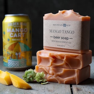 May include: A stack of three orange-hued "Mango Tango" beer soaps with a label, a can of "Mango Cart" beer, mango slices, and hops on a wooden surface. The soap is made with mango wheat ale.