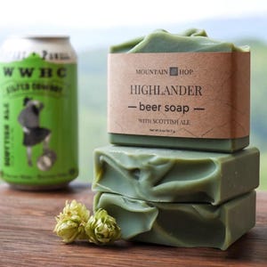 May include: Three green Highlander beer soaps with Scottish ale, stacked on a wooden surface. A can of WWBC Scottish Ale is to the left. Hops are in the foreground. The soap is labeled "Highlander - beer soap".
