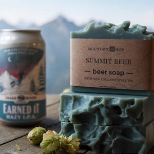 May include: A stack of three green Summit Beer soap bars with a brown label. A can of Earned It Hazy IPA beer is to the left. Hops are scattered around the soap and beer can. The background shows a mountain range.