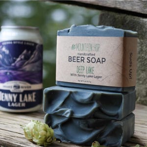 May include: A stack of three blue-grey handcrafted beer soaps labeled "Deep Lake" with "Jenny Lake Lager." A can of "Jenny Lake Lager" and a hop cone are also in the image. The soap is 5 oz / 141.7 g.