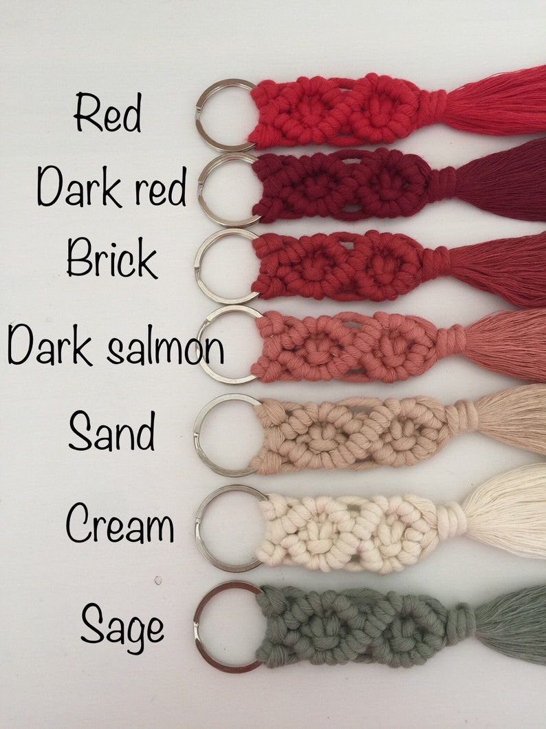 May include: Seven keychains with different colored tassels. The keychains are made of macrame and have a silver key ring. The colors of the tassels are red, dark red, brick, dark salmon, sand, cream, and sage.