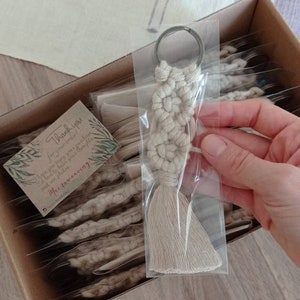 May include: A beige macrame keychain with a silver key ring. The keychain is handmade and has a tassel. The keychain is packaged in a clear plastic bag. The bag is placed in a cardboard box with other keychains. A small card with the text 'Thank you for your order! I hope you enjoy your purchase and look forward to seeing you again soon!  Macrameaning' is visible in the box.