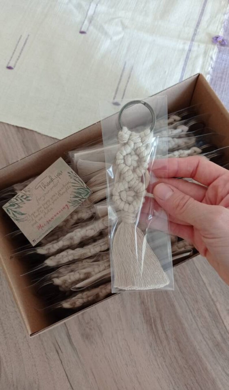 May include: A beige macrame keychain with a silver key ring. The keychain is handmade and has a tassel. The keychain is packaged in a clear plastic bag. The bag is placed in a cardboard box with other keychains. A small card with the text 'Thank you for your order! I hope you enjoy your purchase and look forward to seeing you again soon!  Macrameaning' is visible in the box.