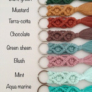 May include: Eight keychains with different colored tassels and macrame designs. The colors are dark green, mustard, terra-cotta, chocolate, green sheen, blush, mint, and aqua marine. The keychains are arranged vertically with the text 'New colors' at the top and the color names listed below each keychain.