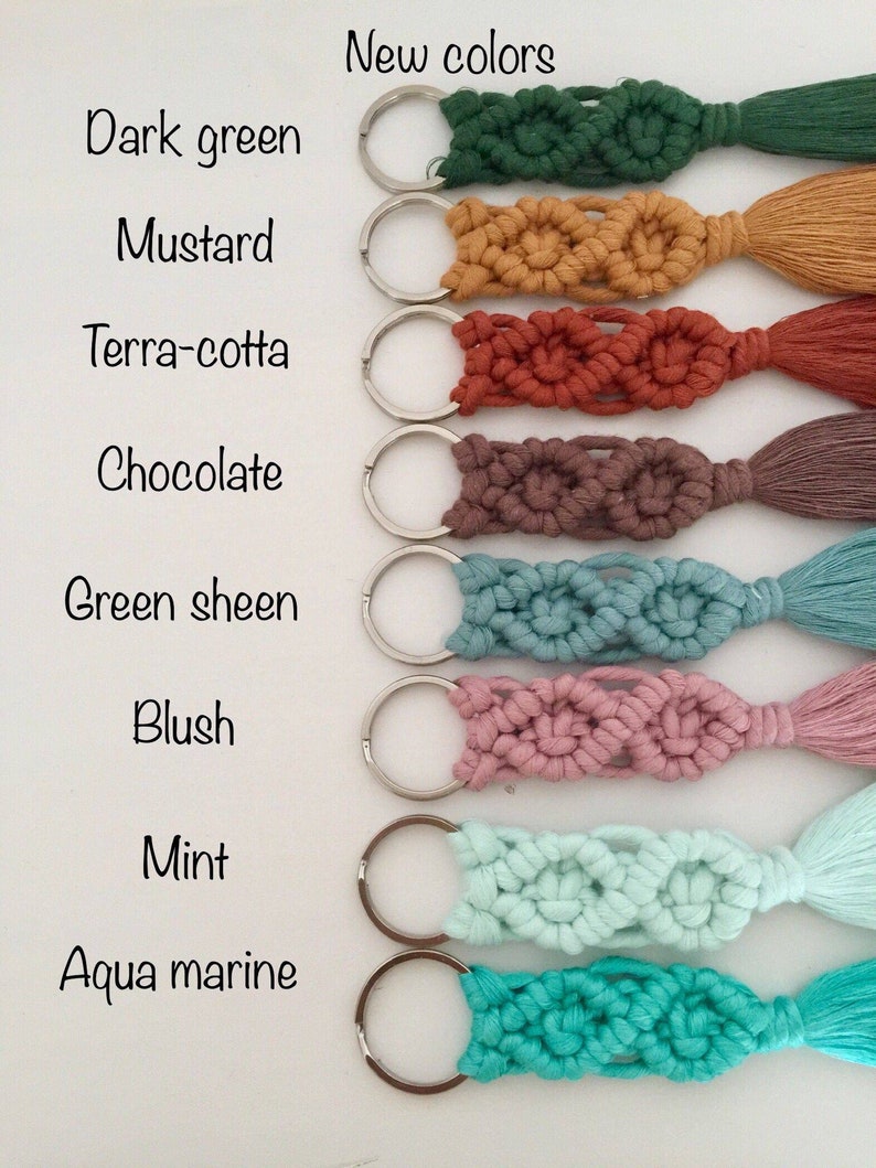 May include: Eight keychains with different colored tassels and macrame designs. The colors are dark green, mustard, terra-cotta, chocolate, green sheen, blush, mint, and aqua marine. The keychains are arranged vertically with the text 'New colors' at the top and the color names listed below each keychain.