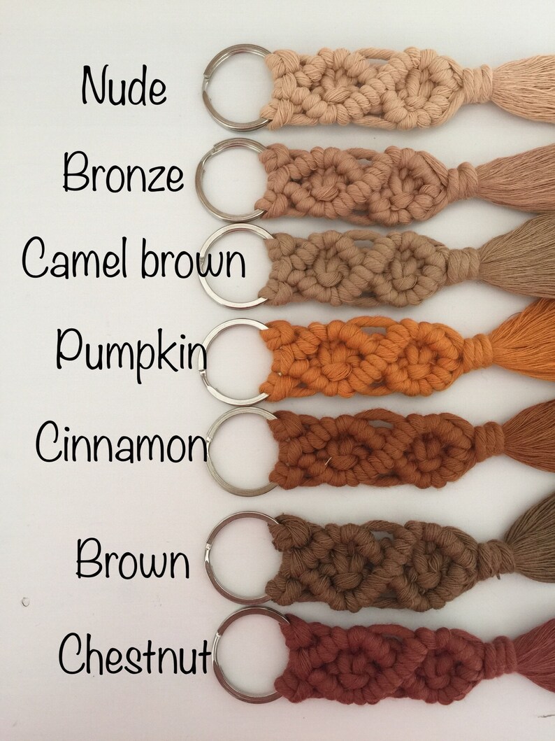 May include: Seven keychains with tassels in different shades of brown, from light to dark. The colors are: Nude, Bronze, Camel Brown, Pumpkin, Cinnamon, Brown, and Chestnut. Each keychain has a silver key ring.