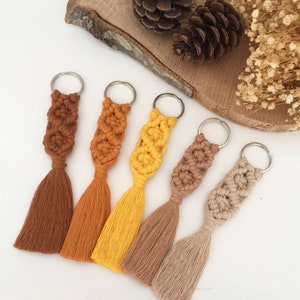 May include: Five keychains with tassels. The keychains are made of macrame and have different colors: brown, orange, yellow, beige, and light beige. The keychains are arranged in a row on a white surface.