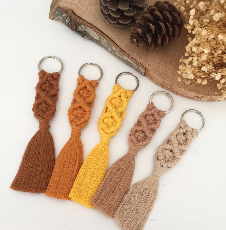 May include: Five keychains with tassels. The keychains are made of macrame and have different colors: brown, orange, yellow, beige, and light beige. The keychains are arranged in a row on a white surface.