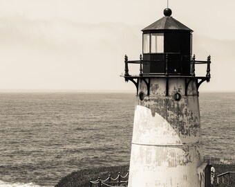 May include: A black and white photo of a lighthouse on a cliff overlooking the ocean. The lighthouse is tall and white with a black roof and a black lantern room. There is a white picket fence in the foreground.