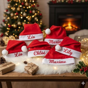 Peut inclure: Plusieurs bonnets de Noël rouges avec bordure blanche et pompons, chacun brodé d'un nom en blanc. Les chapeaux sont disposés sur une table en bois, avec des cadeaux emballés et des décorations festives en arrière-plan.