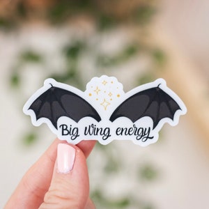 May include: A white sticker with a black silhouette of two wings and gold glitter. The text "Big wing energy" is written below the wings.