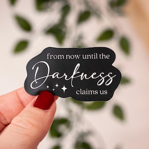 Może przedstawiać: Czarna naklejka z białym napisem: "from now until the Darkness claims us". Naklejka ma unikalny kształt i detale w postaci gwiazdek. Trzymana jest w dłoni z czerwonym lakierem do paznokci.