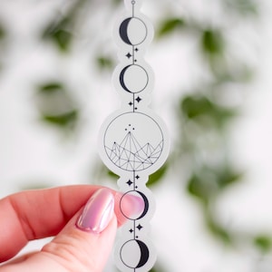 May include: A clear sticker featuring a black and white design of the phases of the moon, stars, and a mountain range. The sticker is held by a hand with a pink iridescent nail polish.