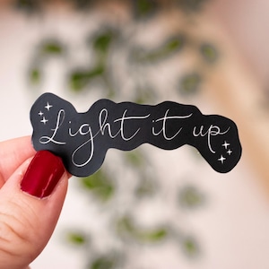 May include: A black sticker with the phrase "Light it up" in elegant white script. The sticker has a wavy shape and is decorated with small starbursts. The sticker is held by a hand with red nail varnish.