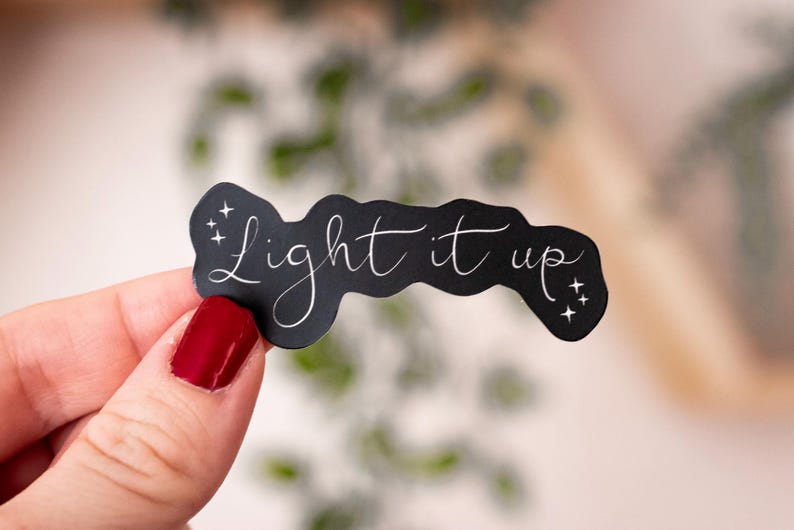 May include: A black sticker with the phrase "Light it up" in elegant white script. The sticker has a wavy shape and is decorated with small starbursts. The sticker is held by a hand with red nail varnish.