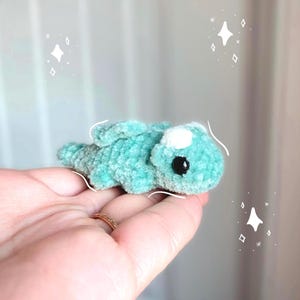 Puede incluir: Un pequeño dragón de ganchillo turquesa con una mancha blanca en la cabeza y ojos negros. El peluche está tumbado, con las alas ligeramente visibles. El dragón está hecho de hilo suave y texturizado.