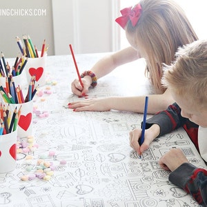 May include: Two children color a large white tablecloth with colorful pencils. The tablecloth has a Valentine's Day theme with hearts and other designs. The pencils are in red cups with hearts on them.