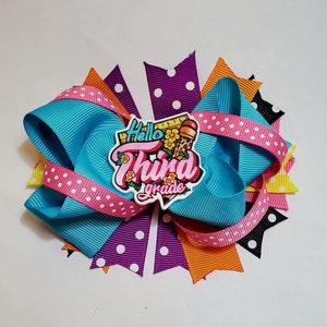 May include: A colorful hair bow with layers of ribbons in turquoise, orange, purple, pink, yellow, and black. The bow features a central emblem that reads "Hello Third Grade" with school-themed graphics.