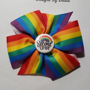 May include: A rainbow-colored hair bow with a white center button that reads "Team Second Grade." The bow is made of colorful, striped ribbon in a fan shape. The text "Designs By Dudas" is written in cursive above the bow.