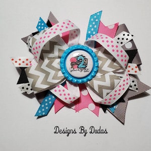May include: A multi-layered hair bow featuring various ribbons in white, pink, gray, black, and turquoise. The bow has a central embellishment with the number "2" and the text "second of the team" on a blue background. The bow is decorated with polka dots and chevron patterns.