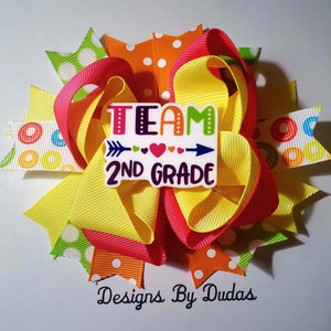 May include: A colorful hair bow with layered ribbons in yellow, orange, and pink. The bow features a white rectangular centerpiece with the words "TEAM 2ND GRADE" and an arrow design. The bow is decorated with polka dots and circular patterns.