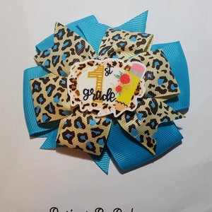May include: A handmade hair bow featuring a turquoise base with layers of tan leopard print ribbon and a central embellishment. The embellishment reads "1st grade" with a pencil and floral accents. The bow is a school-themed accessory.
