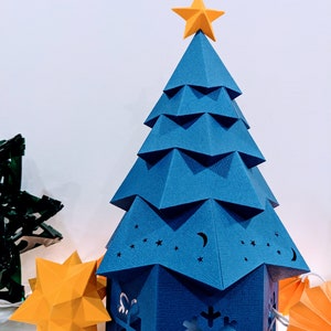 May include: A blue paper Christmas tree with a yellow star on top. The tree has a house-shaped base with cut-out designs of stars, a crescent moon, and a snowflake.