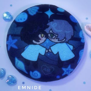 May include: A circular plush pin featuring a dark blue background with a printed illustration of two cartoon characters holding hands. The design includes light blue starfish, seashells, and a heart with the letters AB. The text EMNIDE is visible.