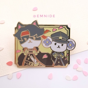 May include: Gold enamel pin featuring two cartoon animals, a black cat with a red bandana and a white bear wearing a blue and gold military uniform. The pin is set against a yellow background with pink hearts and flowers. The text "@EMNIDE" is at the top of the pin.