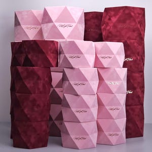 May include: A collection of geometric, velvet-covered boxes in shades of pink and burgundy. Each box features a diamond-like pattern and gold text that reads "Rich Girl Finds". The boxes are stacked in a visually appealing arrangement.