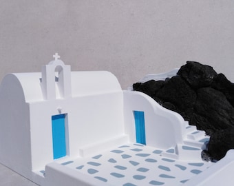 May include: A white model of a Greek church with a blue door and a bell tower. The church is set against a backdrop of black volcanic rocks.