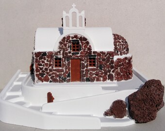 May include: A whitewashed miniature model of a Greek church with a red stone facade and a white dome roof. The church is set on a white base with a small stone path leading up to the entrance.