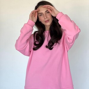 Pode incluir: Um moletom rosa claro com mangas compridas. O moletom é de cor lisa e tem gola redonda. O tecido parece ser um material macio. O moletom faz parte de um conjunto a condizer.