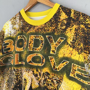 Peut inclure: T-shirt vintage &agrave; col rond avec col jaune et inscription "BODY GLOVE" en grandes lettres soulign&eacute;es. Le t-shirt pr&eacute;sente un motif abstrait noir et jaune, et est suspendu &agrave; un cintre en bois.
