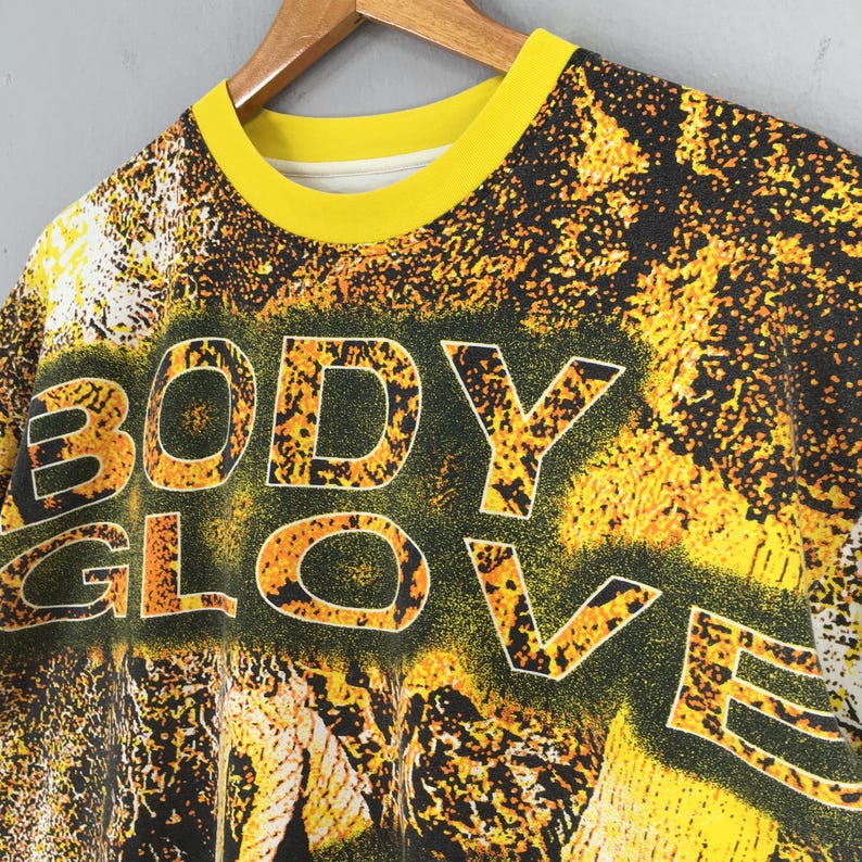 Peut inclure: T-shirt vintage &agrave; col rond avec col jaune et inscription "BODY GLOVE" en grandes lettres soulign&eacute;es. Le t-shirt pr&eacute;sente un motif abstrait noir et jaune, et est suspendu &agrave; un cintre en bois.