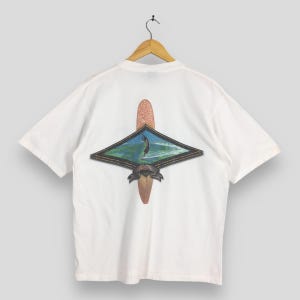May include: White t-shirt with a surfboard graphic on the back. The graphic features a surfer riding a wave inside a diamond-shaped frame, with two surfboards. The shirt is hanging on a wooden hanger.
