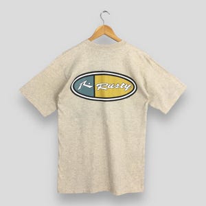 May include: A light gray t-shirt with a blue and yellow oval logo that says "Rusty" on the back.